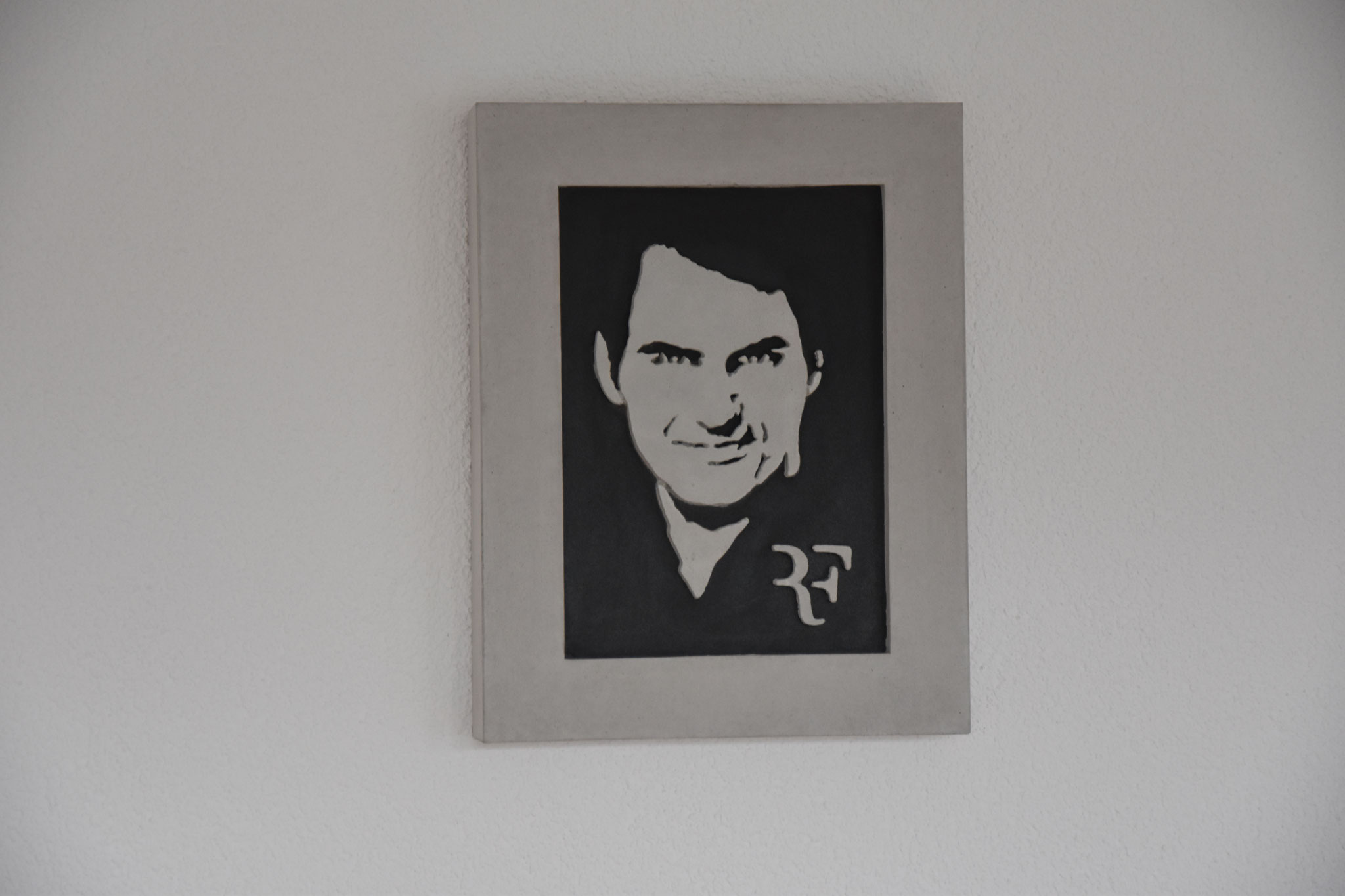 Betonportrait von Roger Federer mit seinem ikonischen Logo ‚RF‘, dargestellt auf schwarzem Hintergrund und umrahmt von Beton.